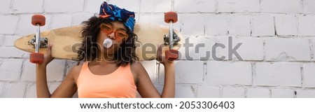 stylish african american woman in sunglasses and headscarf blowing bubble gum and holding longboard near brick wall, banner