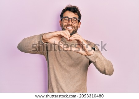 Handsome man with beard wearing turtleneck sweater and glasses smiling in love doing heart symbol shape with hands. romantic concept. 