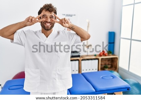 Young handsome physiotherapist man working at pain recovery clinic doing peace symbol with fingers over face, smiling cheerful showing victory 