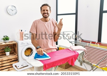 Young hispanic man ironing clothes at home gesturing finger crossed smiling with hope and eyes closed. luck and superstitious concept. 