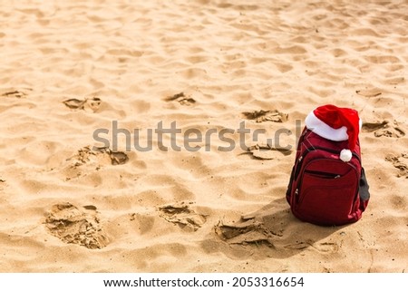Santa hat on a tourist red backpack against the blue sea, enjoying the sunset of the ocean horizon. Christmas beach vacation travel banner or card