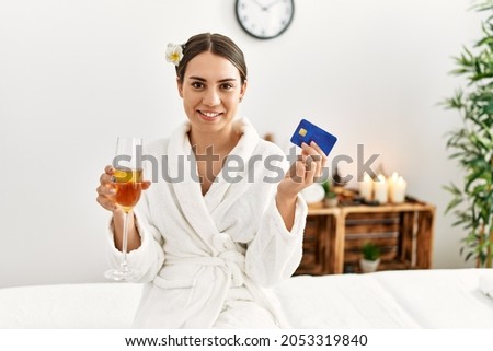 Young hispanic woman holding credit card drinking champagne at beauty center.