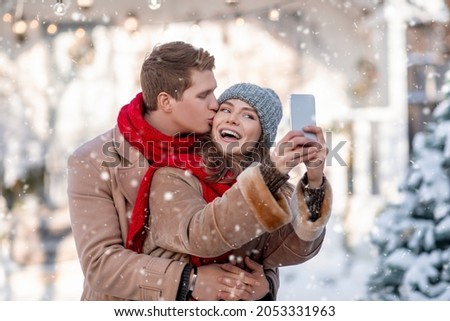 Cheerful young couple enjoying first snow at park, taking selfie together, using smartphone. Loving guy kissing his happy girlfriend while she is taking photo on cellphone, white backyard background