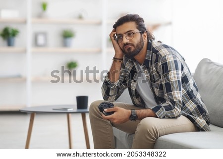 Bored indian guy sitting on couch with joystick, looking at copy space, playing video games alone at home. Middle-eastern young man feeling lonely while spending weekend alone, home entertainment Royalty-Free Stock Photo #2053348322