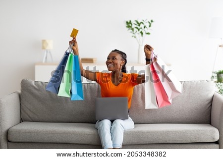 Joyful black woman sitting on sofa with laptop and credit card, purchasing in online shop, buying on sale from home. Excited female customer with gift bags using electronic store service Royalty-Free Stock Photo #2053348382