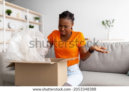 Terrible delivery service, online shopping. Unhappy irritated black woman unboxing cardboard parcel, receiving damaged item at home. African American lady feeling disappointed about wrong parcel Royalty-Free Stock Photo #2053348388