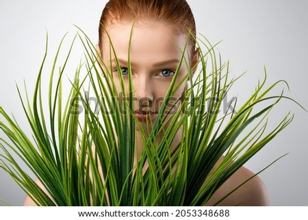 Natural Female Beauty. Red-Haired Girl Posing With Plants Looking At Camera Through Green Grass On Gray Studio Background. Wellness And Spa, Cosmetics Advertisement Concept