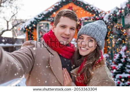 Cheerful lovers young man and woman taking selfie while spending snowy winter day together outside, loving millennial couple celebrating winter holidays, taking photo at backyard while snowing