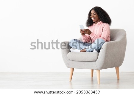 Cheerful african american young lady sitting in arm chair at home, using nice mobile application on brand new smartphone, shopping online, white background, panorama with copy space Royalty-Free Stock Photo #2053363439