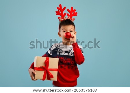 Holiday Fun. Cheerful Asian Boy Wearing Deer Horns And Ugly Sweater With Ornament Holding Christmas Present, Playful Kid Keeping Xmas Tree Ball Near Nose And Looking At Camera, Blue Background