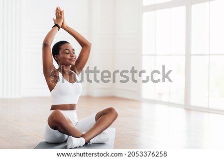 Young black sporty woman practicing yoga, doing Sukhasana exercise, Easy Seat pose raising arms up lifting hands, working out near fitness studio window, wearing white pants and top, free copy space Royalty-Free Stock Photo #2053376258