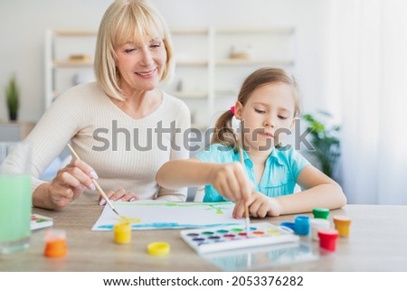 Children Creativity Concept. Portrait of little girl painting with watercolors and gouache at home together with granny. Happe mature lady helping grandchild creating picture, sitting at table