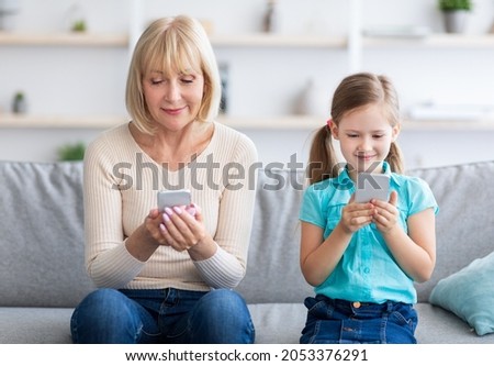 Modern Lifestyle, Family Gadgets. Portrait of smiling mature woman and little preschool kid girl using two smart phones sitting separate on the couch in living room and looking at screens