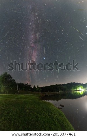 Panorama of the night sky with the Milky Way and the meteor swarm Perseids 2021. In the foreground a lake and a meadow on the shore. 