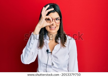 Young hispanic woman using lavalier microphone smiling happy doing ok sign with hand on eye looking through fingers 