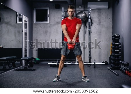 A confident attitude and a seductive look of a muscular sportsman performing exercises with a kettle bell. Dead lifts and squats with a load in the indoor space of a modern gym. Royalty-Free Stock Photo #2053391582