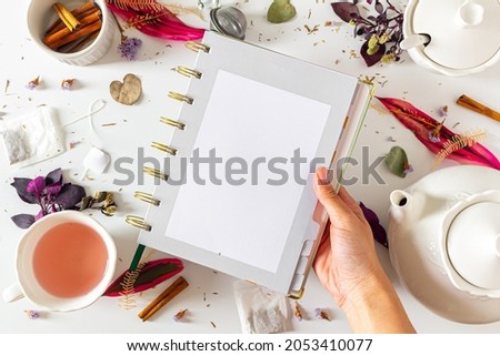 Tea time femine frame composition with female hand holding a planner, tea pot, cinnamon, cup, tea strainer, purple and pink leaves and dried plants  on white background. Flat lay, top view. 