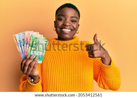 Young african american woman holding south african rand banknotes smiling happy and positive, thumb up doing excellent and approval sign  Royalty-Free Stock Photo #2053412561