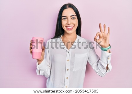 Beautiful woman with blue eyes drinking a take away cup of coffee doing ok sign with fingers, smiling friendly gesturing excellent symbol 