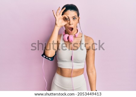Young hispanic woman wearing gym clothes and using headphones doing ok gesture shocked with surprised face, eye looking through fingers. unbelieving expression. 