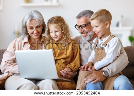 Smiling senior grandparents and cute little grandchildren boy and girl looking at laptop computer screen while sitting on sofa at home, happy kids watching cartoons with grandpa and grandma on weekend