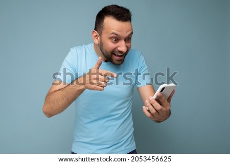 Emotional handsome young bearded brunet man wearing everyday blue t-shirt isolated over blue background holding and using mobile phone communication online on the internet looking at gadjet display