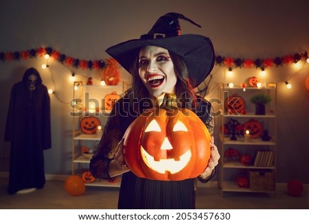 Horror night. Halloween portrait of spooky and evil witch with glowing jack-o-lantern in hands. Young woman in witch's hat with crazy and creepy smile looks into camera while standing in dark room.