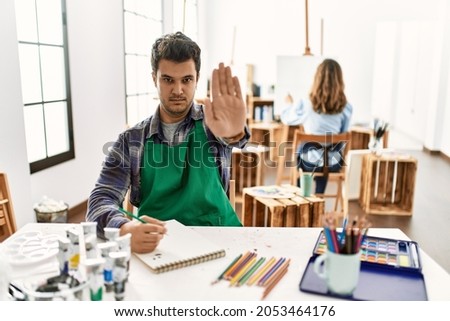 Young artist man at art studio doing stop sing with palm of the hand. warning expression with negative and serious gesture on the face. 
