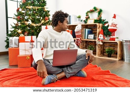 Young african american man using laptop sitting by christmas tree looking to side, relax profile pose with natural face with confident smile. 