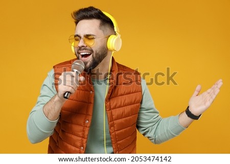 Young singer man 20s years old wearing orange vest mint sweatshirt eyeglasses headphones singing song in microphone spreading hand in karaoke club isolated on yellow color background studio portrait.