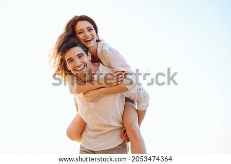 Young playful fun happy enamored smiling couple two friends family man woman in casual clothes boyfriend give piggyback ride to joyful girlfriend sit on back look aside on light white sky background Royalty-Free Stock Photo #2053474364