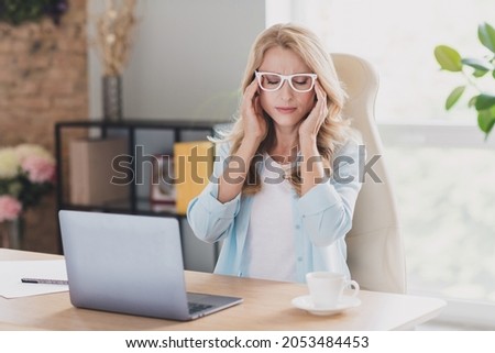 Photo of tired upset mature lady wear blue shirt glasses sitting table typing modern device arms temples indoors house room