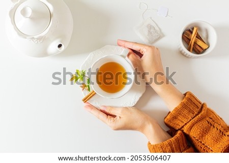 Composition with female hands holding a cup of tea, teapot, cinnamon and tea bag on white background. Flat lay, top view. Slow morning concept. Royalty-Free Stock Photo #2053506407