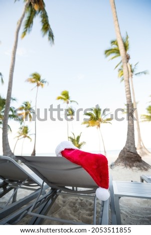 Santa hat hanging on chaise lounge with coconut palm trees and bounty beach scene. Travel and New Year holiday card. 