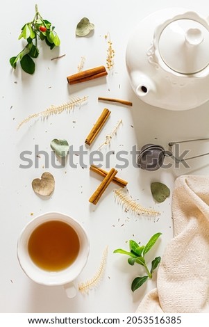 Composition with tea pot, cup  of tea, cinnamon, tea strainer, linen fabric, green plant leaves on white background. Flat lay, top view. 