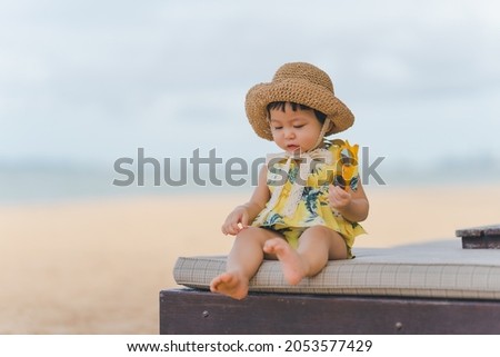 A baby with moments of excitement and joy to be out at sea. by sitting on a beach chair and wearing a hat and holding sunglasses and wearing a bright yellow dress