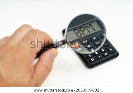 Business and finance concept. A man looks through a magnifying glass at a calculator on the screen of which it is written - BPM Royalty-Free Stock Photo #2053590602