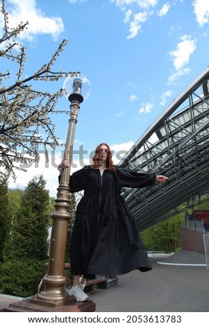 Model showing autumn clothes. Ginger emotionless girl in oversize summer long wide black raincoat dress hanging on lights pole. Performing, dancing and urban culture concept. Fashion concept.