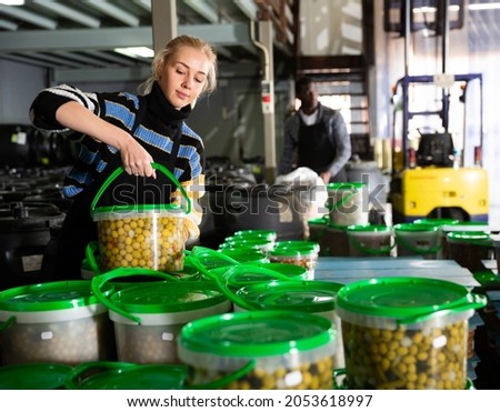 Female worker stocks plastic containers and cans with olives in warehouse. High quality photo