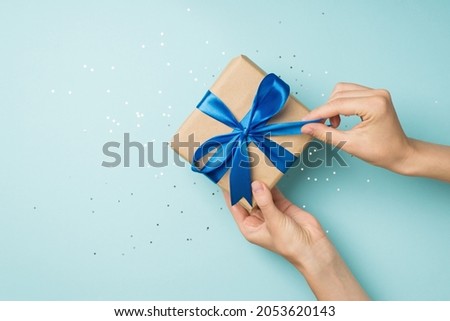 First person top view photo of hands unpacking craft paper gift box with blue satin ribbon bow over shiny sequins on isolated pastel blue background with blank space Royalty-Free Stock Photo #2053620143