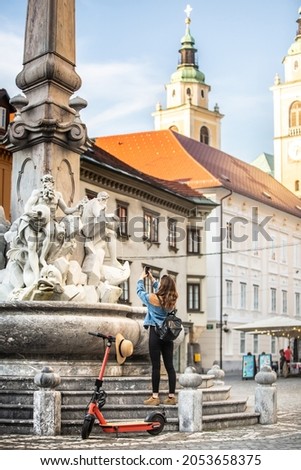 Traveler exploring old town on environmentally friendly electric scooter. Female tourist exploring Ljubljana's old medieval historical city center, taking picture of a fountain with her phone. Royalty-Free Stock Photo #2053658375