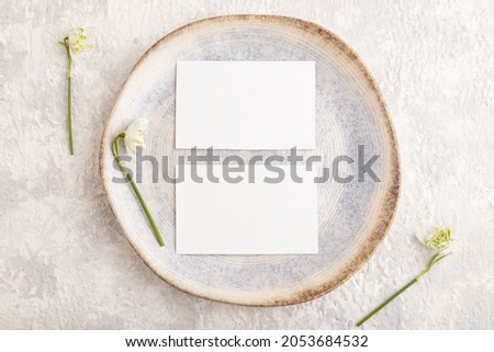 White paper invitation card, mockup with galanthus snowdrop flowers on ceramic plate and gray concrete background. Blank, flat lay, top view, still life, copy space, wedding invitation.