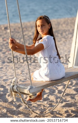 A cute brunette girl 10 years old in a white dress swinging on a swing on the seashore in summer.