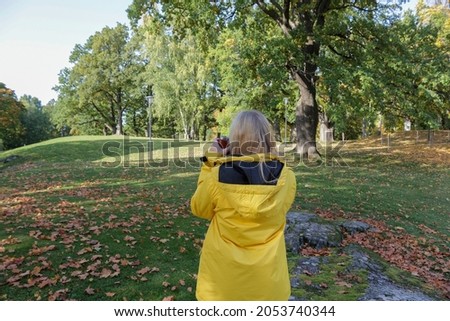 A teenage girl in a yellow jacket takes pictures of the city park on the camera. Hobby photography