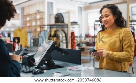 Clothing Store Checkout Cashier Counter: Woman Retail Sales Manager Accept NFC Smartphone and Credit Card Payments from a Young Female Customers for Clothes. Royalty-Free Stock Photo #2053746086