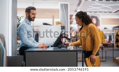 Clothing Store: Young Woman at Counter Buys Clothes from Friendly Retail Sales Assistant, Paying with Contactless NFC Smartphone Touching Terminal. Trendy Fashion Shop with Designer Brands. Royalty-Free Stock Photo #2053746191