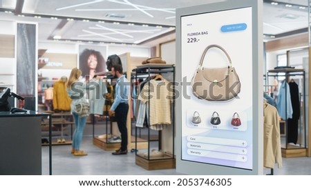 Shot of a Floor-Standing LCD Touch Screen Display with User Interface of Online Clothing Shop Standing in Clothing Store. Self service Checkout with Hand Bag. Diverse People in Shop Buying Clothes. Royalty-Free Stock Photo #2053746305