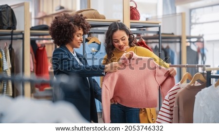 Young Female Customer Shopping in Clothing Store, Retail Sales Associate Helps with Advice. Diverse People in Fashionable Shop, Choosing Stylish Clothes, Colorful Brand with Sustainable Designs Royalty-Free Stock Photo #2053746332