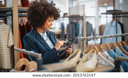 Clothing Store: Female Merchandising Manager Uses Tablet Computer To Create Stylish Fashion Collection. Professional Shop Sales Retail Assistant Checks Stock. Small Business Owner Orders Mall Items. Royalty-Free Stock Photo #2053763147