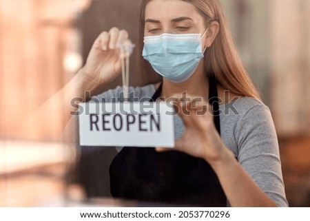 small business, reopening and service concept - woman in mask hanging reopen banner on window or door glass